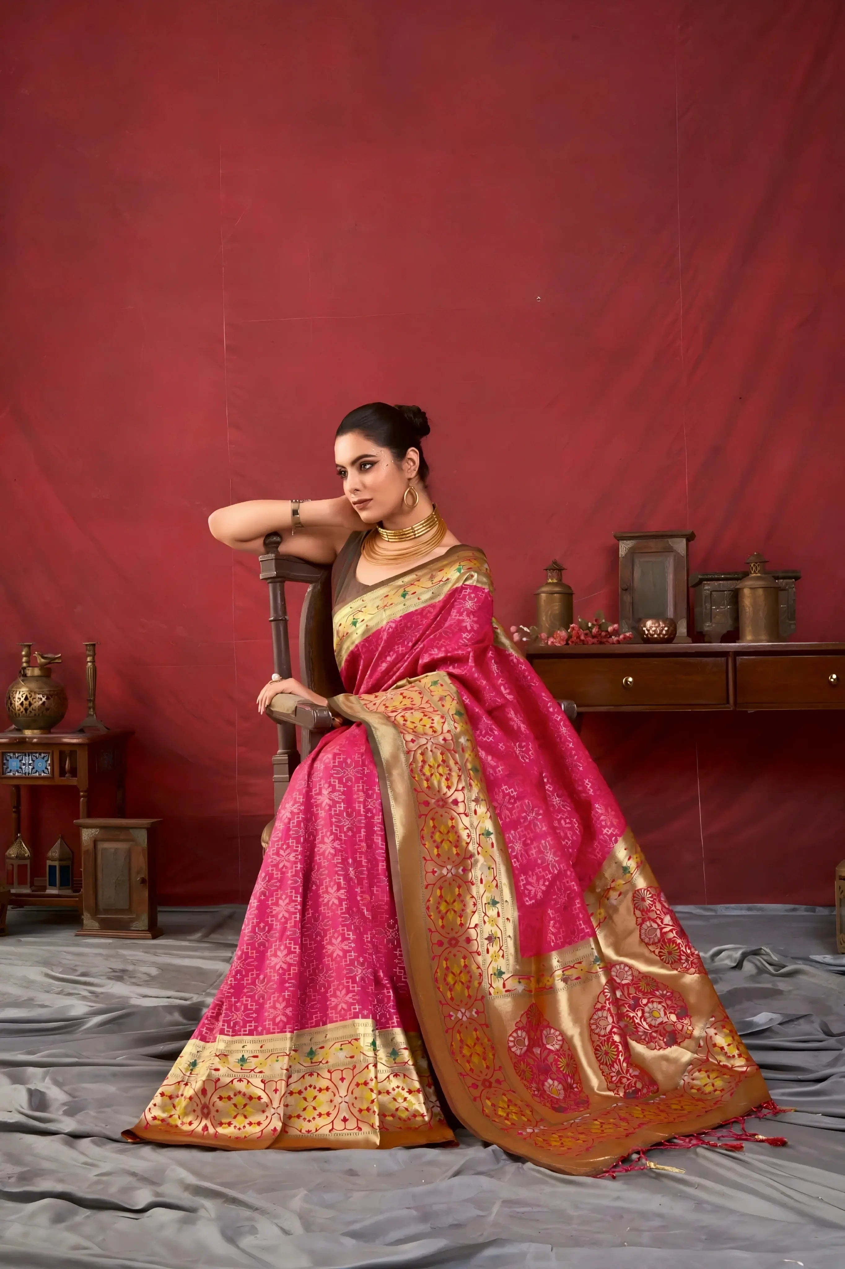 A woman models a luxurious Hot Pink Soft Banarasi Silk Saree. The saree features dense, all-over tone-on-tone Zari weaving. The wide border and pallu showcase heavy gold Zari work with vibrant green and yellow accents. She wears a contrasting dark (black/brown) sleeveless blouse and a heavy gold Kundan choker necklace, posing indoors.